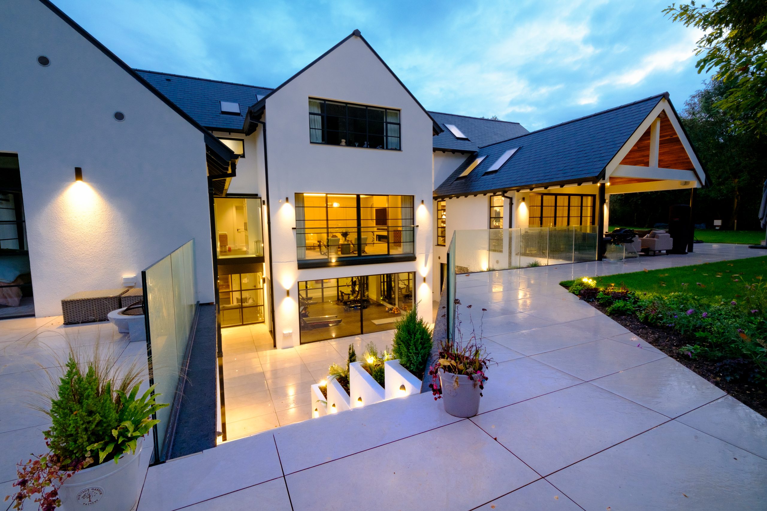 The back side of a luxury house with the garden, a lower basement terrace with glass balustrades, and steel look windows with bespoke glazing bars
